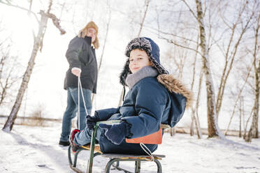 Lächelnder Junge, der auf einem Schlitten sitzend im Winter über die Schulter schaut - NLAF00283