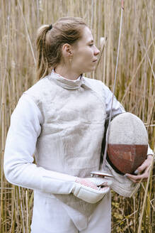 Fencer holding mask and foil standing amidst grass - YHF00109