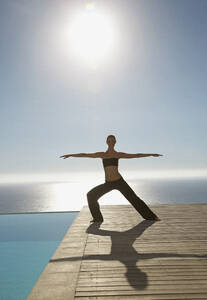 Junge Frau übt Yoga an einem Swimmingpool mit dem Meer im Hintergrund - FSIF06814