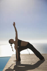 Junge Frau übt Yoga an einem Swimmingpool mit dem Meer im Hintergrund - FSIF06816