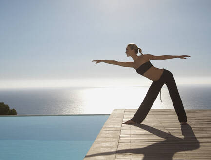 Junge Frau übt Yoga an einem Swimmingpool mit dem Meer im Hintergrund - FSIF06819