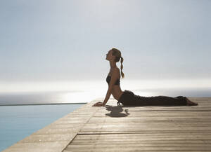Junge Frau übt Yoga an einem Swimmingpool mit dem Meer im Hintergrund - FSIF06821