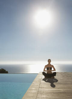 Junge Frau übt Yoga an einem Swimmingpool mit dem Meer im Hintergrund - FSIF06822