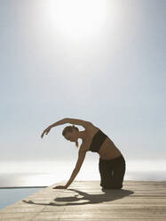 Junge Frau übt Yoga an einem Swimmingpool mit dem Meer im Hintergrund - FSIF06823