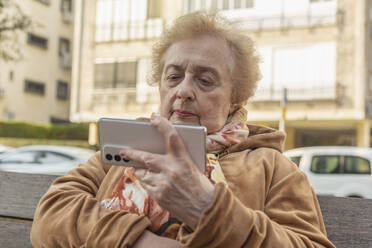 Ältere Frau sitzt und benutzt ihr Smartphone auf einer Bank in der Stadt - FSIF06877