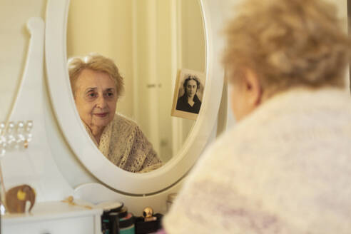 Lächelnde ältere Frau betrachtet ihr Spiegelbild neben einem alten Foto - FSIF06878