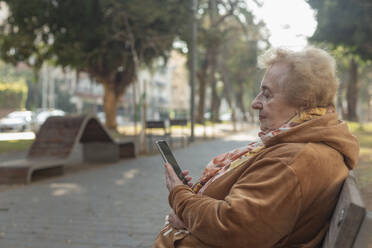 Ältere Frau sitzt und benutzt ihr Smartphone auf einer Parkbank in der Stadt - FSIF06880