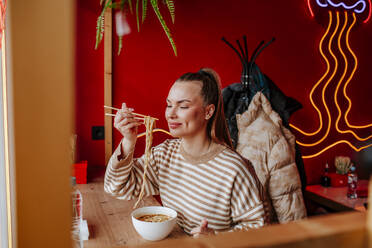 Smiling woman with eyes closed holding ramen noodles in chopsticks at restaurant - MDOF01822