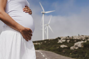 Schwangere Frau mit Händen auf dem Bauch in der Nähe von Windkraftanlagen - EBBF08592