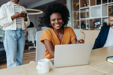 Glückliche Unternehmerin mit Brille arbeitet mit Kollegen an einem modernen Arbeitsplatz zusammen. Reife Geschäftsfrau, die in die Kamera schaut, während sie während einer kreativen Besprechung mit einem Laptop sitzt. - JLPSF31175