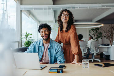 Two professionals, a man and a woman, collaborating in a modern office. They smile, looking at the camera. A creative workspace represents teamwork, diversity, and success. - JLPSF31182