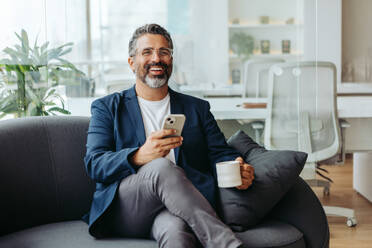 Glücklicher, reifer Geschäftsmann, der auf einem grauen Sofa sitzt, eine Tasse Kaffee in der Hand hält und eine SMS auf seinem Smartphone schreibt. Älterer Unternehmer in legerer Geschäftskleidung in einem professionellen Büro. - JLPSF31190