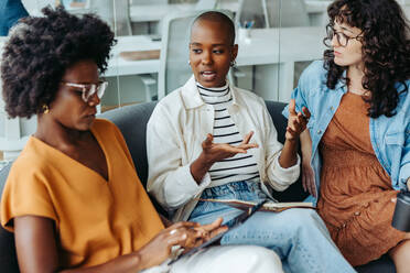 Eine Gruppe verschiedener Geschäftsfrauen beim Brainstorming auf einer modernen Bürocouch. Lächelnd, fröhlich und professionell zeigen sie Teamwork und Kreativität an einem Startup-Arbeitsplatz. - JLPSF31195