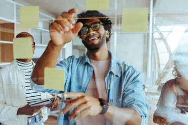 Business professionals engage in a collaborative meeting using a glass wall, full of creative ideas and positive energy. The team uses sticky notes for strategic planning, showcasing their effective teamwork and leadership. - JLPSF31200