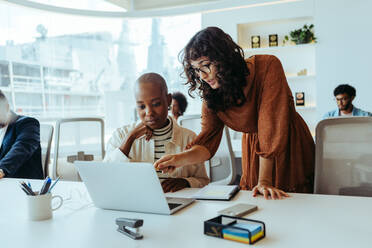 Zwei Geschäftsfrauen, die in einem modernen Büro zusammenarbeiten, einen Laptop benutzen und über ein Projekt diskutieren. Sie sind glücklich und lächeln, was ihre Teamarbeit an einem technologiegesteuerten Arbeitsplatz zeigt. - JLPSF31213