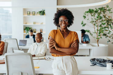 Confident black woman with afro hair and glasses stands in a vibrant office. She is an entrepreneur and businesswoman, smiling and looking at the camera with crossed arms. Her modern business casual attire reflects her creativity in the workplace. - JLPSF31228