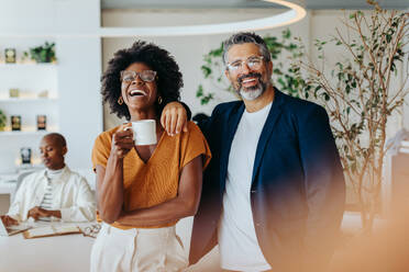 Zwei Geschäftsleute stehen in einem Büro und halten Kaffeetassen in der Hand. Sie sind glücklich und lächeln, was Teamwork und Erfolg widerspiegelt. Geschäftsmann und Geschäftsfrau zeigen Professionalität und Vertrauen. - JLPSF31233