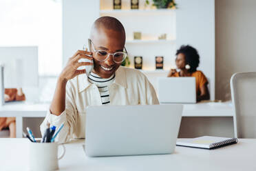 Eine glückliche, junge Geschäftsfrau führt ein Telefongespräch, während sie in einem modernen Büro an ihrem Laptop arbeitet. Im Hintergrund ist ein Kollege zu sehen, der sich auf seine Arbeit konzentriert. - JLPSF31239