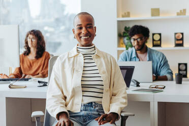 Smiling black woman with glasses sits in an office. She is a successful entrepreneur surrounded by colleagues in a startup workplace. They all look at the camera, showcasing their business casual attire. - JLPSF31240