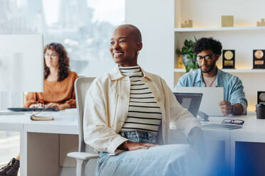 Eine selbstbewusste schwarze Unternehmerin sitzt in einem modernen Büro mit Geschäftskollegen. Sie trägt eine Brille, lächelt und blickt nachdenklich in die Ferne. Sie arbeiten gemeinsam in einem Startup und strahlen eine positive, fröhliche Atmosphäre aus. - JLPSF31241