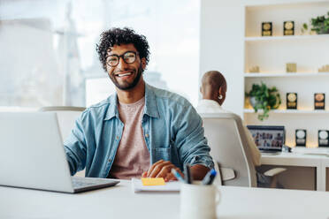 Young male entrepreneur with curly hair and glasses sits at a modern office table. He is working on a laptop and appears happy and smiling. The professional is surrounded by technology and is successful in his startup business. - JLPSF31243