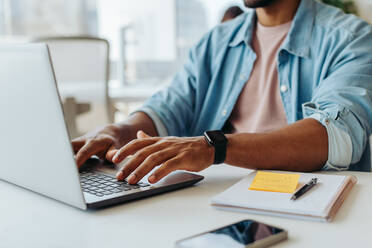 Geschäftsmann sitzt an einem modernen Bürotisch und tippt auf einem Laptop. Junger männlicher Unternehmer, der die Technologie zu seinem Vorteil nutzt und sich darauf konzentriert, in seinem Start-up-Unternehmen erfolgreich zu sein. - JLPSF31252