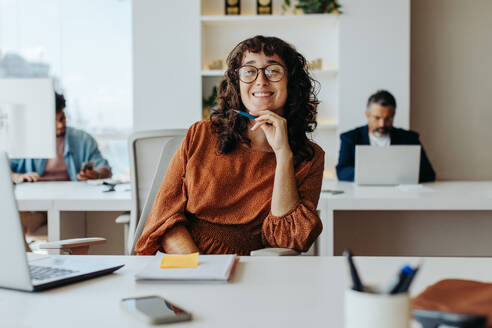 Erfolgreiche Geschäftsfrau, die an einem Tisch in ihrem Büro arbeitet, eine Brille trägt und lächelt. Sie ist auf ihr Projekt konzentriert, umgeben von Notizen und zeigt kreativen und unternehmerischen Geist. - JLPSF31254