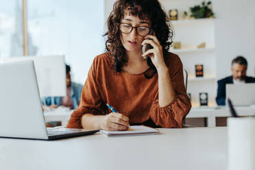 Professionelle Geschäftsfrau mit Brille, die an ihrem Büroschreibtisch sitzt und an ihrem Laptop arbeitet. Sie führt ein Telefongespräch und demonstriert damit ihre Kommunikationsfähigkeiten. - JLPSF31255