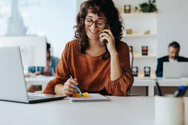 Eine gelockte Geschäftsfrau mit Brille sitzt an einem Tisch in einem Büro und arbeitet fröhlich an ihrem Laptop und ihrem Smartphone. Sie nimmt einen Anruf entgegen und plant ihre berufliche Arbeit, umgeben von Technik. - JLPSF31256