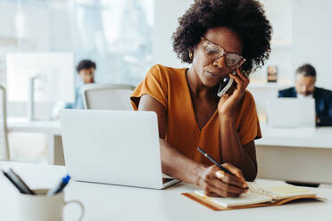 Geschäftsfrau an einem modernen Bürotisch, die einen Laptop und ein Mobiltelefon zur Kommunikation und Planung nutzt. Sie ist konzentriert und engagiert und zeigt eine kreative und professionelle Arbeitsumgebung. - JLPSF31259