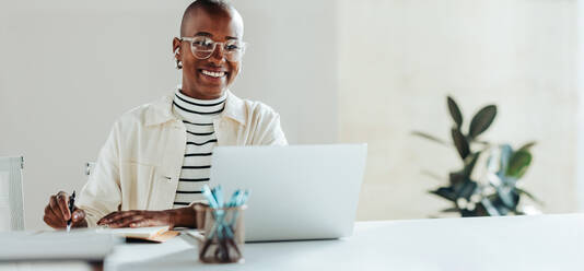 Unternehmerin mit Brille bei der Arbeit im Heimbüro, lächelnd, während sie sich neben einem Laptop Notizen macht. Moderner Arbeitsplatz mit natürlichem Licht und Zimmerpflanzen, der Produktivität und Glück vermittelt. - JLPSF31283