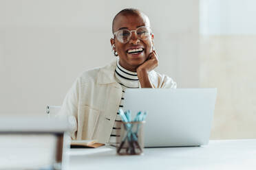 Cheerful black businesswoman with glasses working on a laptop at a bright modern office, exuding positivity and job satisfaction. Casual professional, creative workspace, happy employee. - JLPSF31284