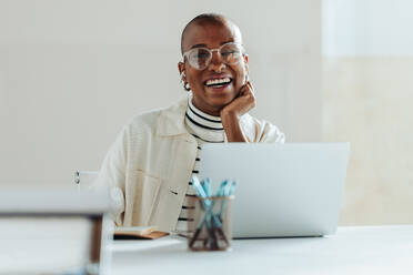 Fröhliche schwarze Geschäftsfrau mit Brille, die in einem hellen, modernen Büro an einem Laptop arbeitet und Positivität und Arbeitszufriedenheit ausstrahlt. Lässiger professioneller, kreativer Arbeitsplatz, glücklicher Mitarbeiter. - JLPSF31284