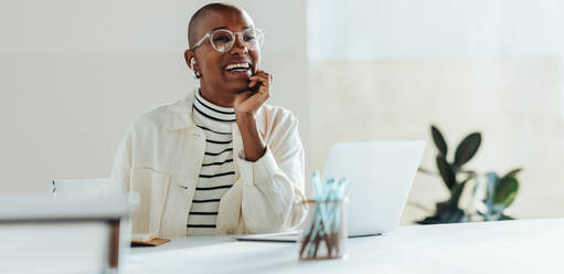 Happy young businesswoman celebrates a successful moment while working on her laptop in a bright, contemporary office setting, showcasing workplace positivity and achievement. - JLPSF31285