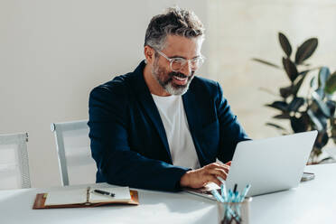 Ein fröhlicher, reifer Geschäftsmann mit Bart arbeitet aufmerksam an seinem Laptop an einem aufgeräumten Schreibtisch in einem modernen Büro und strahlt Zufriedenheit, Professionalität und Produktivität aus. - JLPSF31289