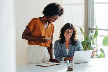 Fröhliche, berufstätige Frauen, die in einem modernen Büro mit Begeisterung zusammenarbeiten. Zwei Unternehmerinnen, die einen Laptop für eine produktive Geschäftsbesprechung in einem Unternehmen nutzen. - JLPSF31291