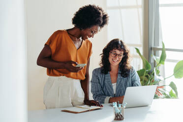 Happy professional women collaborating with enthusiasm in a modern office setting. Two female entrepreneurs using a laptop for a productive business meeting in a corporate workplace. - JLPSF31291