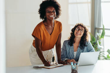 Zwei Frauen mit unterschiedlichen Berufen lächeln in die Kamera, während sie in einem kreativen Büro zusammenarbeiten. Sie strahlen Positivität und Teamwork aus, auf dem Schreibtisch stehen ein Laptop und Büromaterial. - JLPSF31293