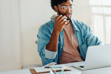 Ein junger, eleganter Geschäftsmann arbeitet in einem hellen, modernen Büro fleißig an seinem Laptop, führt ein Telefongespräch und hält ein Smartphone in der Hand, das Professionalität und Konzentration verkörpert. - JLPSF31304