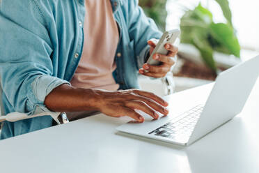 Junger professioneller Mann, der in einem modernen Büro Multitasking betreibt und dabei sowohl ein Smartphone als auch einen Laptop benutzt. Das Bild erinnert an Konzepte von Konnektivität, Arbeitseffizienz und Technologie im Geschäftsleben. - JLPSF31309