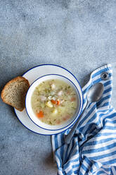 Von oben warme Schüssel mit Hühner- und Gemüsesuppe mit Vollkornbrot auf einem strukturierten grauen Hintergrund mit gestreifter Serviette und einem Löffel - ADSF52765