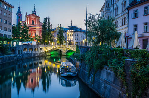 Blick in der Dämmerung auf einen ruhigen Fluss, der durch Ljubljana fließt, mit beleuchteten historischen Gebäuden und einer Brücke, die sich im Wasser spiegelt - ADSF52772