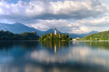 Serene waters of Lake Bled reflect the idyllic church island and castle amidst the Julian Alps on a cloudy day - ADSF52777