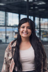 Stylish young curvy female in top and jacket smiling while standing near modern glass office building on street looking at camera - ADSF52802