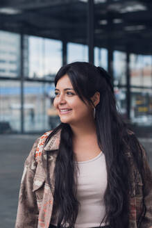 Stilvolle junge kurvige Frau in Top und Jacke lächelt, während sie in der Nähe eines modernen Glasbürogebäudes auf der Straße steht und wegschaut - ADSF52804