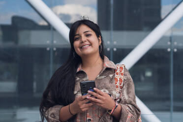 Positive Frau in Armband und Freizeitkleidung lächelnd und mit Smartphone gegen Glasgebäude auf sonnigen Tag - ADSF52806