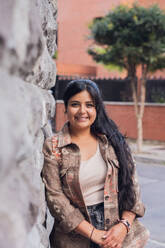 Positive young lady in top and jacket smiling and standing near brick wall and looking at camera on sunny day - ADSF52809