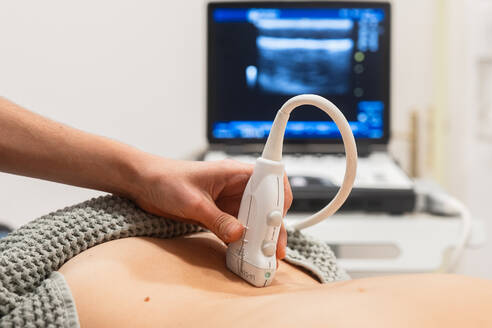 A close-up image of a diagnostic medical ultrasound being performed on a patient's abdomen in a clinical setting, with the ultrasound screen visible in the background - ADSF52835