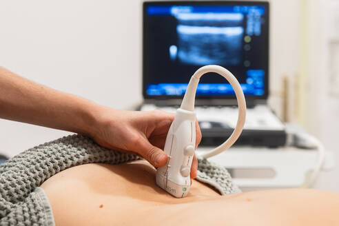 Nahaufnahme eines diagnostischen medizinischen Ultraschalls, der in einer klinischen Umgebung am Bauch eines Patienten durchgeführt wird, wobei der Ultraschallbildschirm im Hintergrund zu sehen ist - ADSF52835