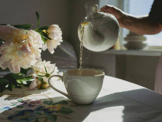 Frau gießt Tee in Tasse in der Nähe von Blumen auf dem Tisch zu Hause - IEF00526